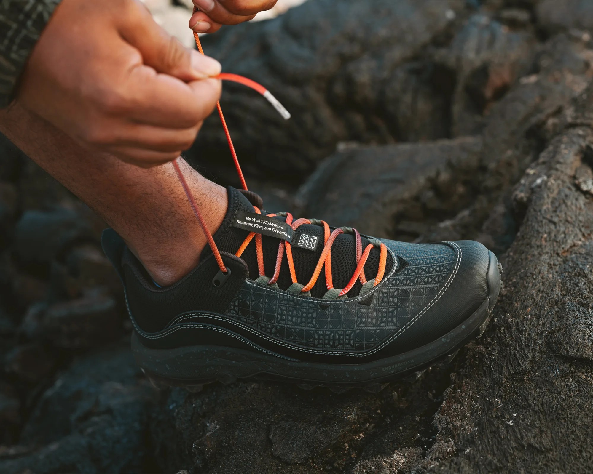 Black and gray hiking shoe with orange laces being tied on a rocky surface.