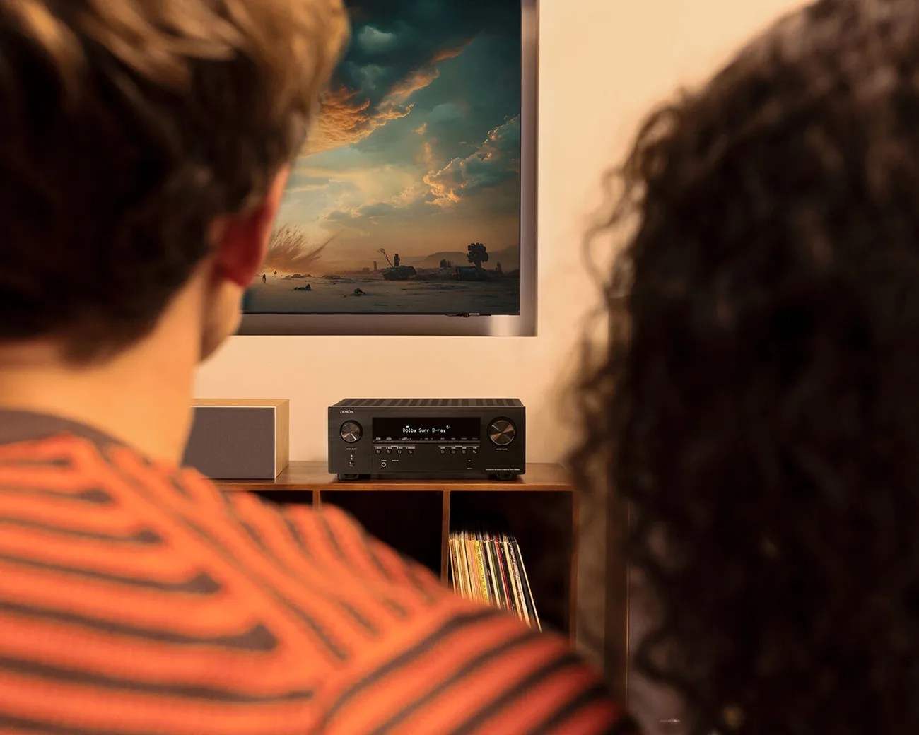 Black Denon audio receiver with display and control knobs on a wooden shelf below a wall-mounted TV.
