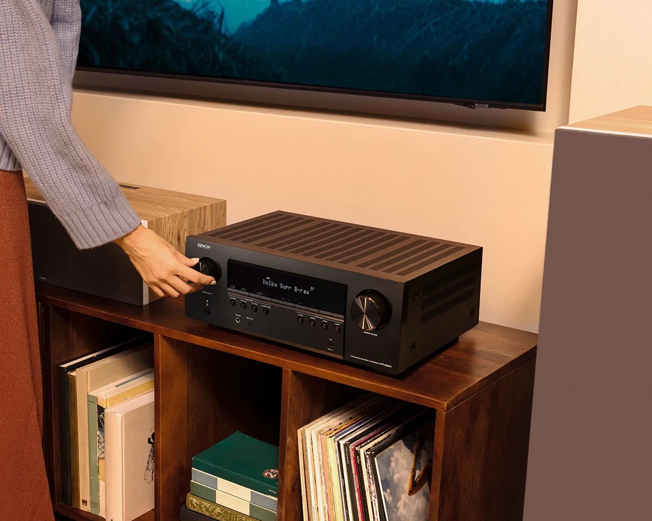 Black Denon audio receiver with knobs and buttons on a wooden shelf, a hand adjusting the left knob.