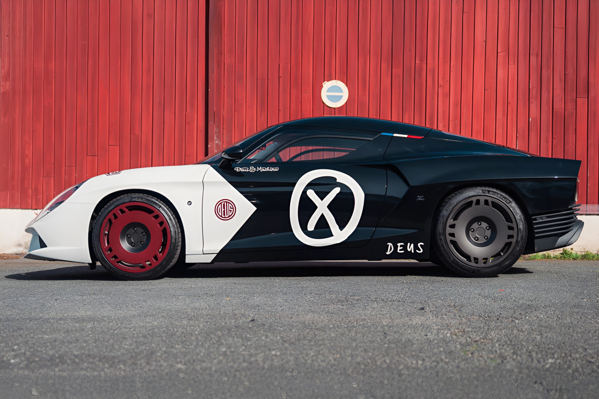 Black and white sports car with red front wheel and large white X logo on the door, parked in front of a red wooden wall.