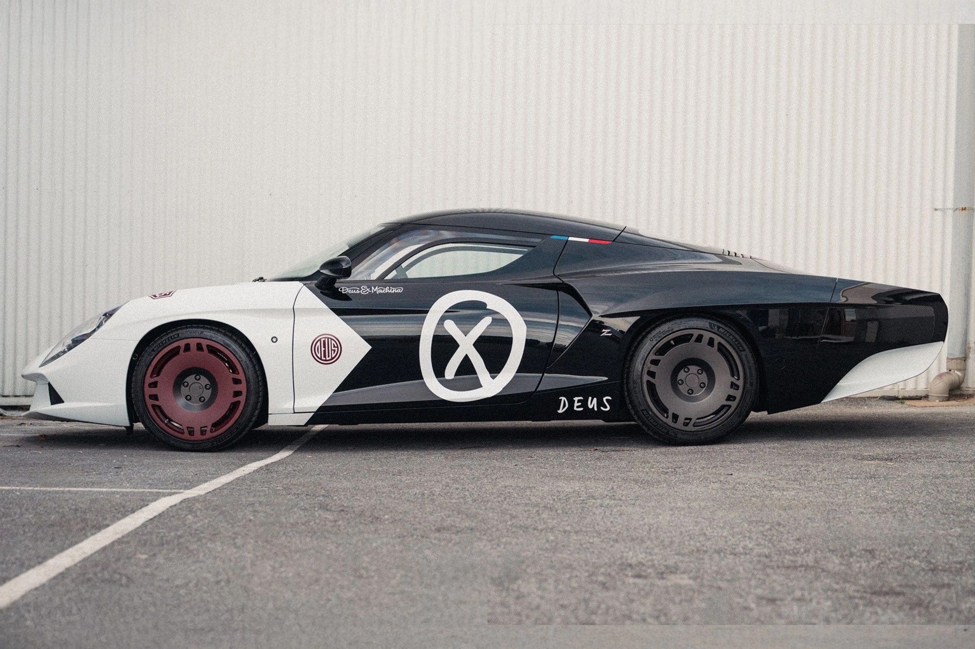 Black and white sports car with red front wheel and black rear wheel, featuring Deus & Machina logos on the side.