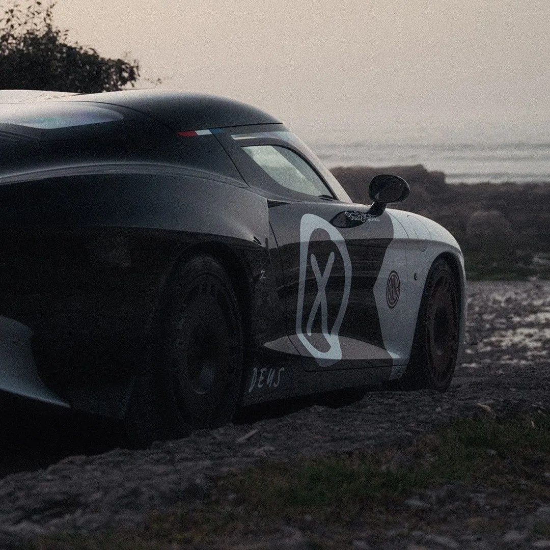 Black and white race car with large "X" logo on the door parked on rocky ground near water at dusk.