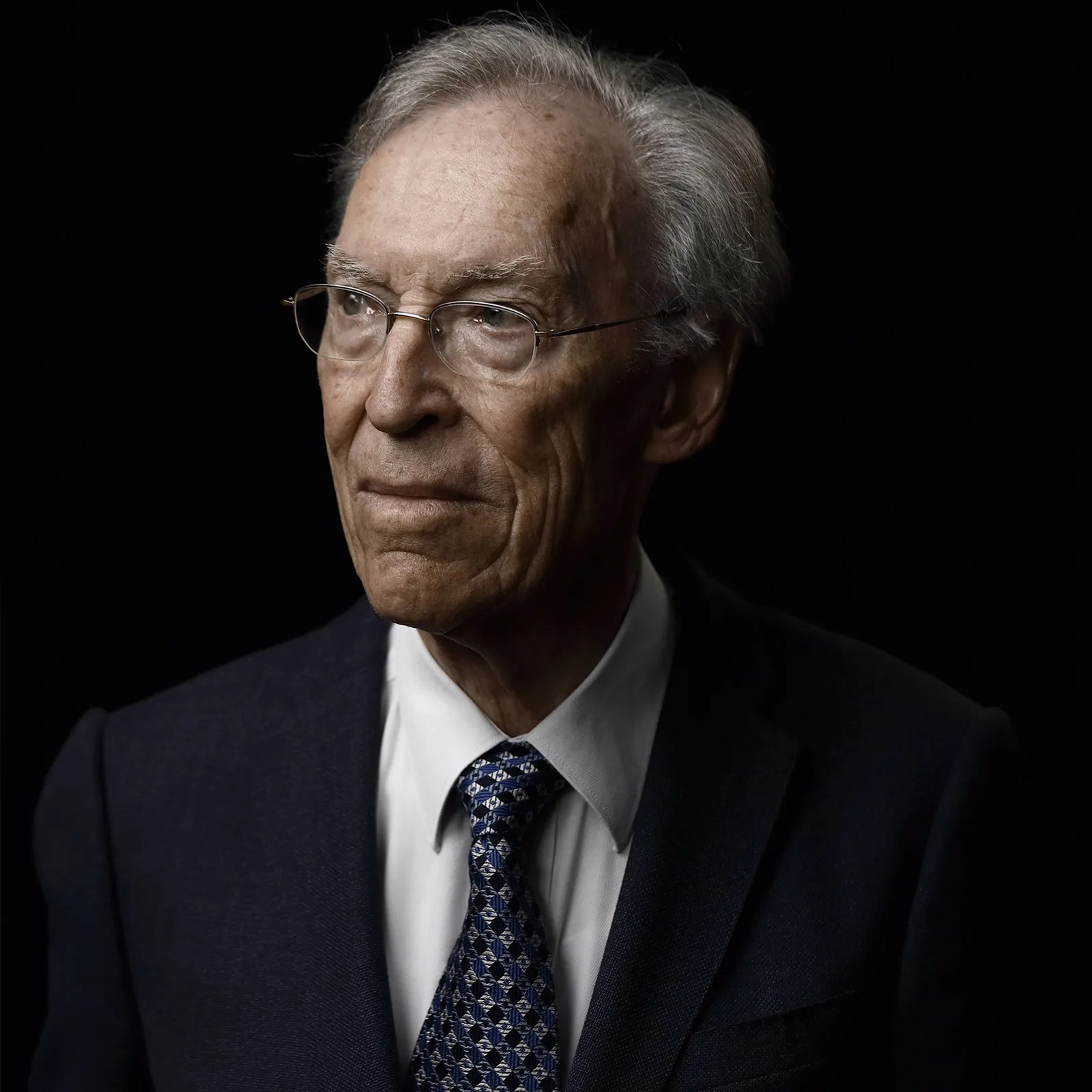 Elderly man with gray hair and glasses wearing a dark suit, white shirt, and patterned blue tie against a black background.