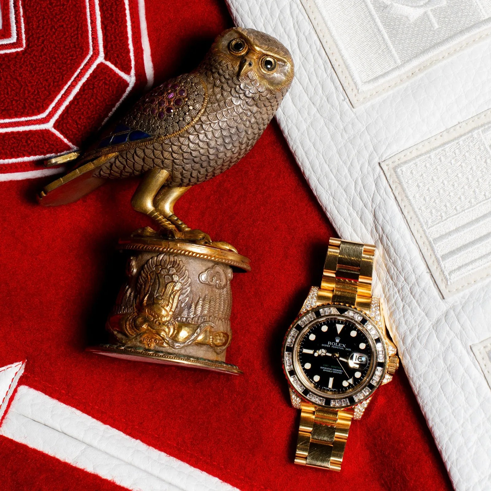 Ornate bronze owl figurine with detailed feathers and a gold Rolex watch with black dial on red and white fabric.