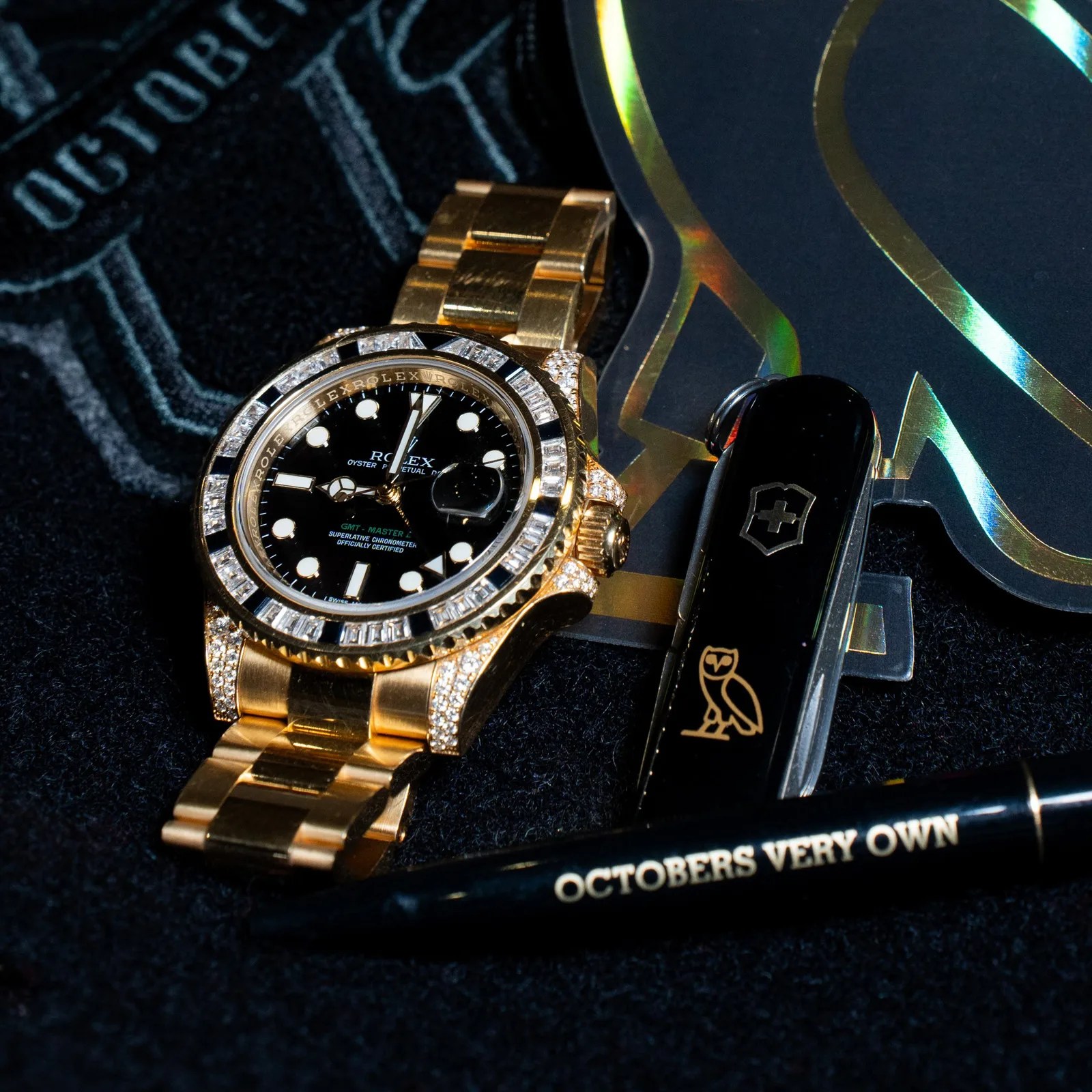 Gold Rolex GMT-Master II watch with diamond bezel and black dial next to a black Swiss Army knife with owl logo and "OCTOBERS VERY OWN" pen on black fabric.