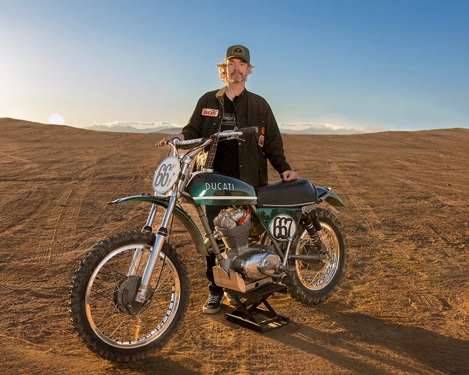 Man standing behind a vintage green Ducati dirt bike with number 667 on a dirt track at sunset.