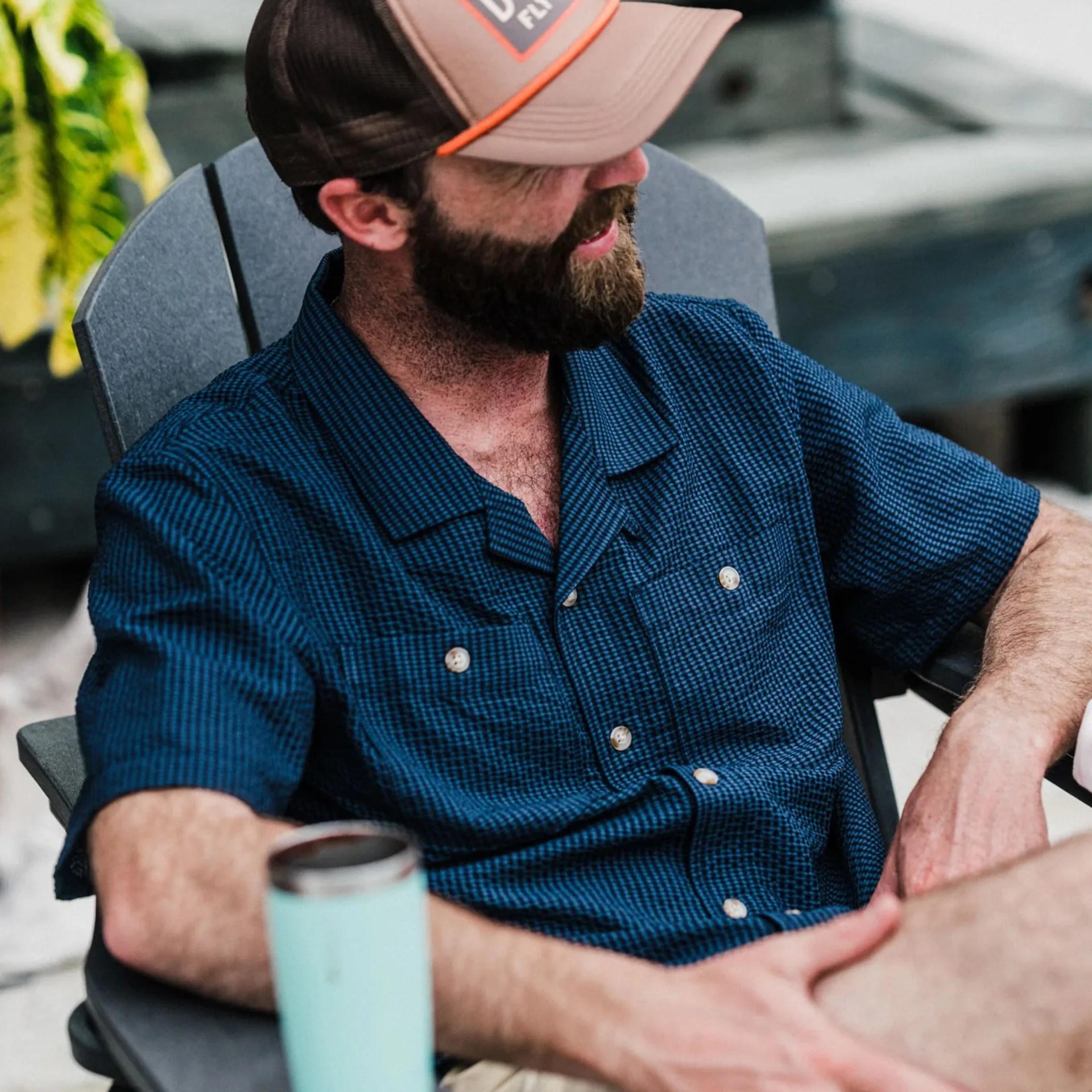 Man wearing a blue checkered short-sleeve button-up shirt and a brown cap sitting in a chair holding a light blue tumbler.