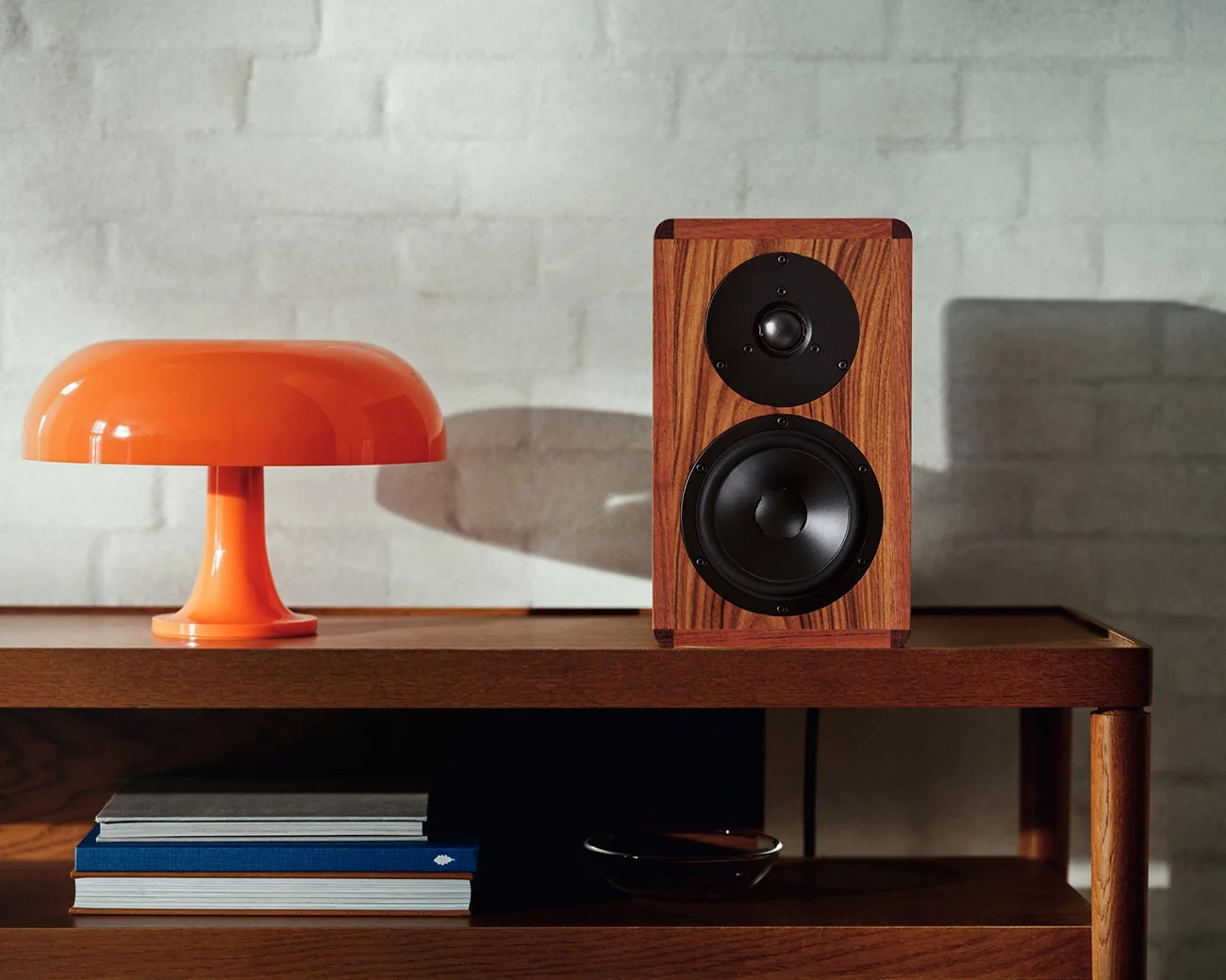 Orange mushroom-shaped table lamp and wooden speaker with black drivers on a wooden shelf against a light brick wall.
