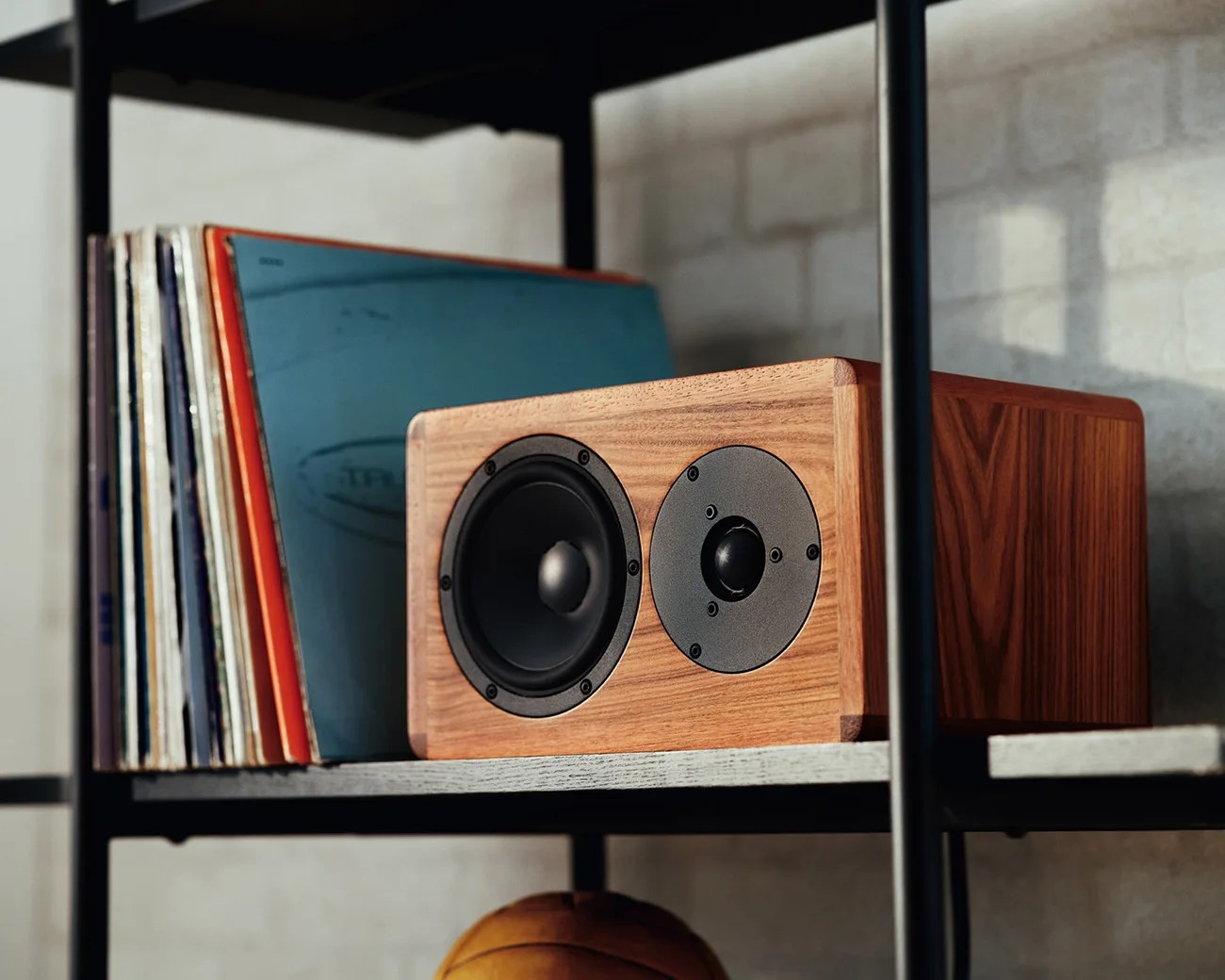 Wooden bookshelf speaker with black drivers next to vinyl records on a gray shelf.