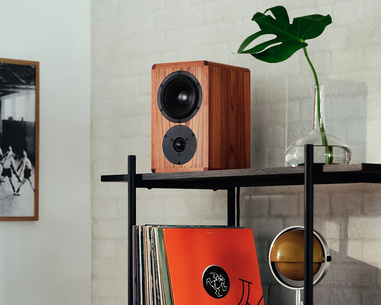 Wooden bookshelf speaker with black drivers on a dark wood and metal shelf next to a glass vase with a green leaf.