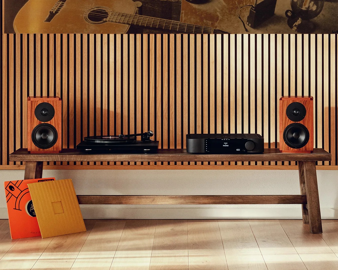 Pair of wooden bookshelf speakers, black turntable, and black audio receiver on a wooden bench against a vertical slat wood wall.