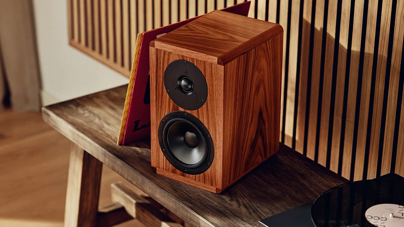Wooden bookshelf speaker with black drivers on a dark wooden bench next to a vinyl record and album cover.
