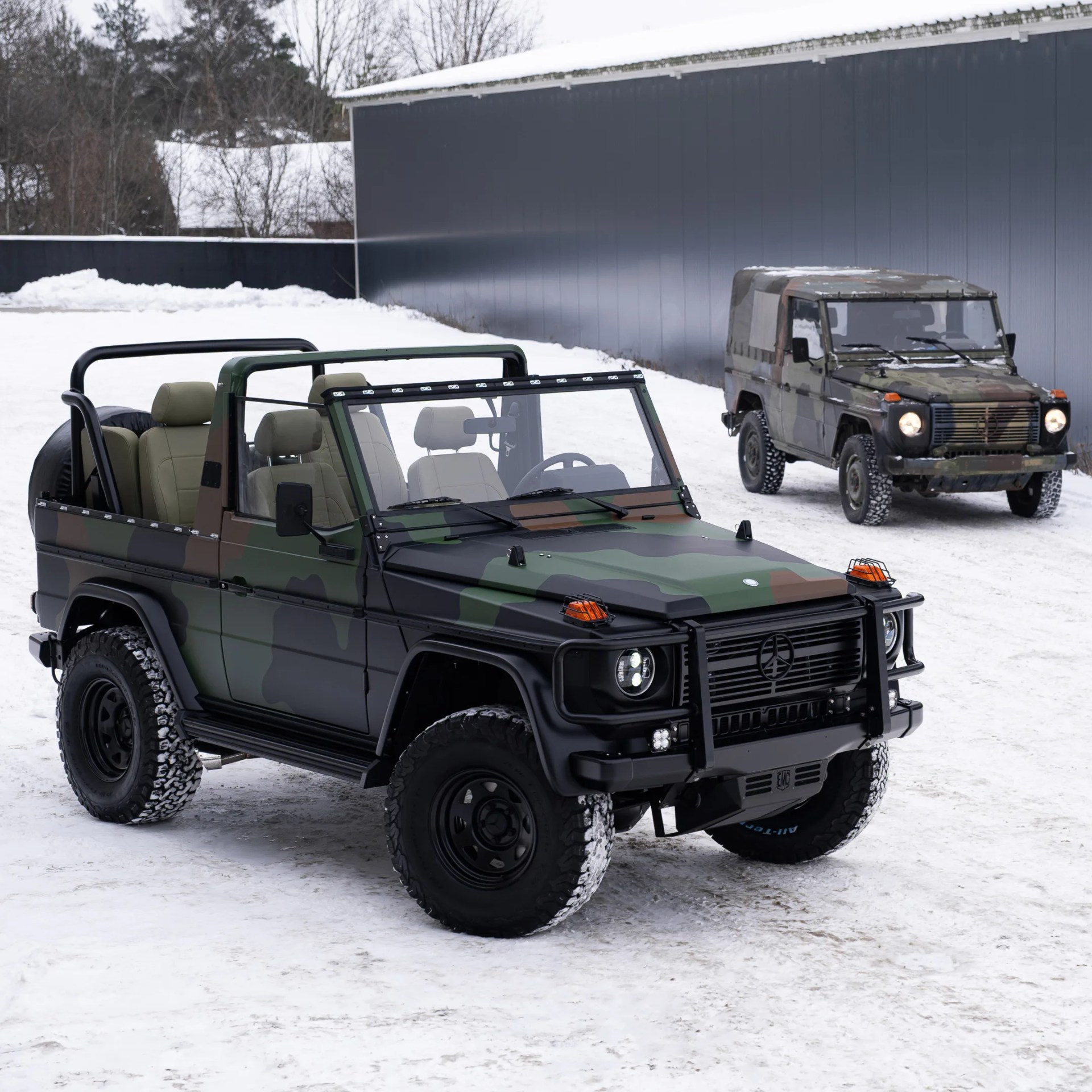 Two camouflaged off-road SUVs with roll bars parked on snow near a large gray building.