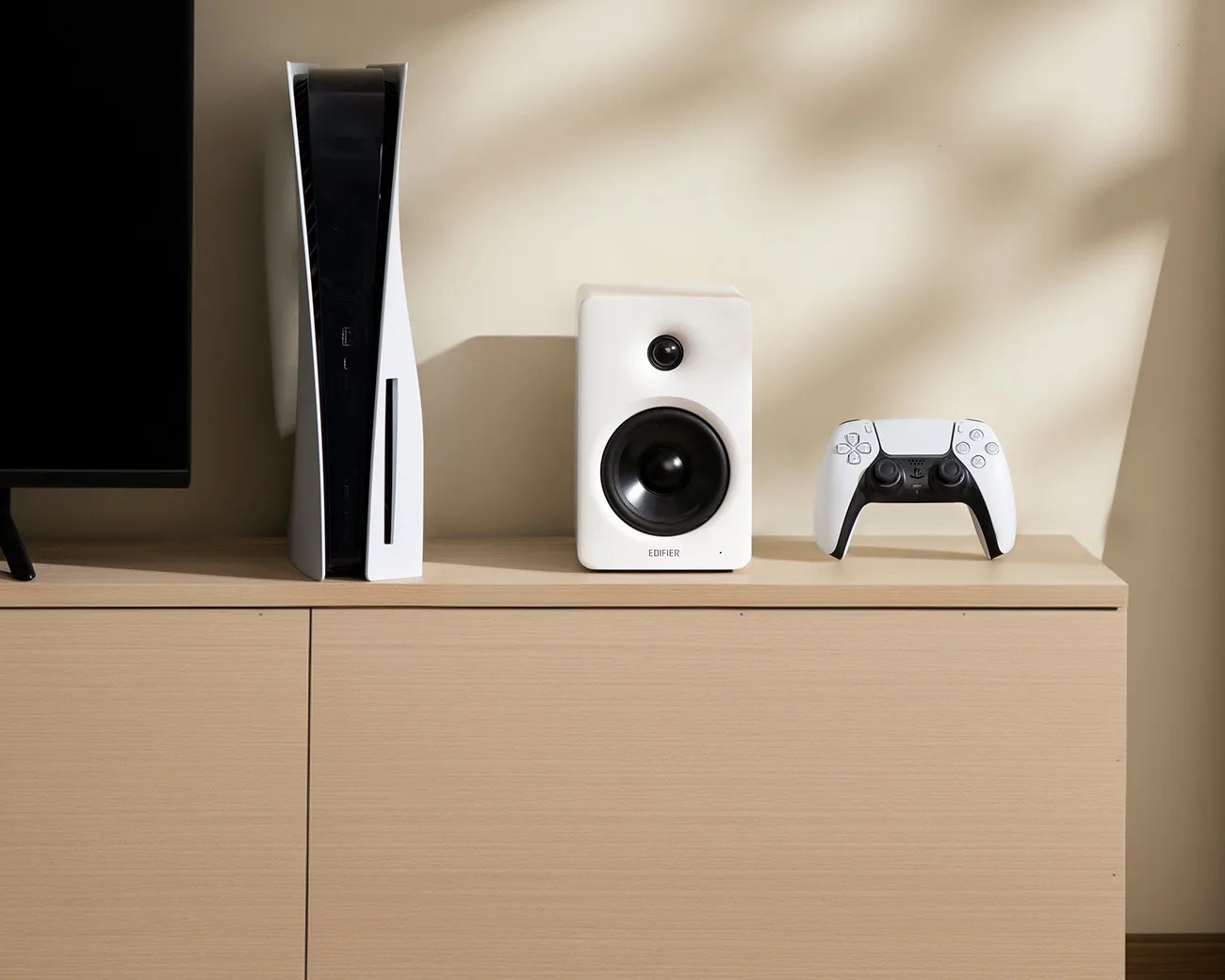 White and black PlayStation 5 console, white Edifier speaker, and white DualSense controller on a light wood cabinet.