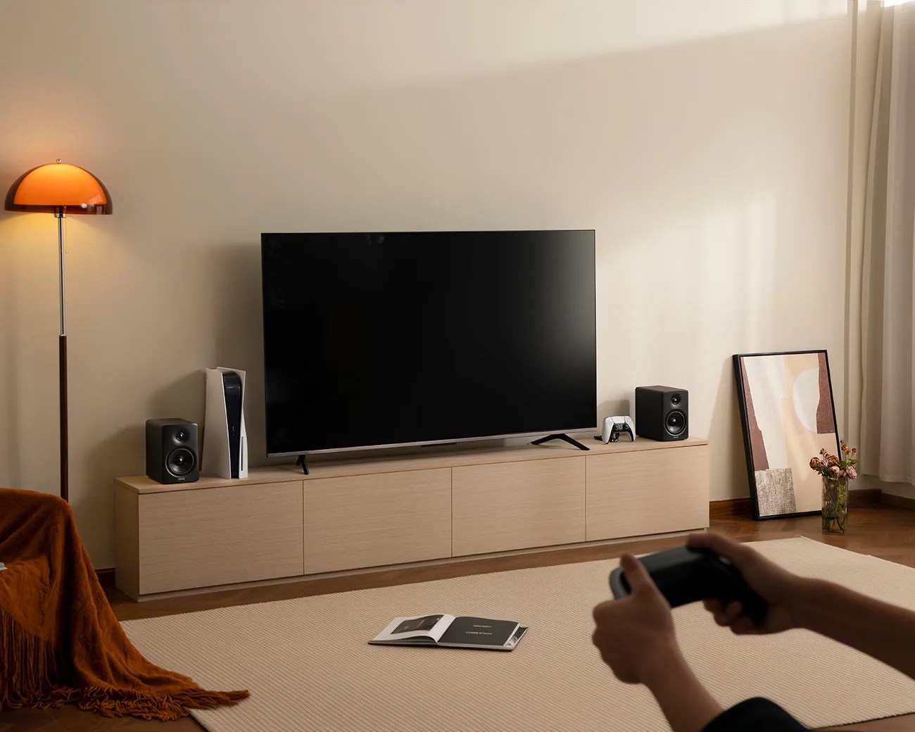 Modern living room with a flat-screen TV on a light wood cabinet, gaming console, speakers, and a person holding a game controller.