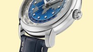 Close-up of a silver watch with a blue and gray world map dial, city names on the bezel, and a dark blue leather strap.