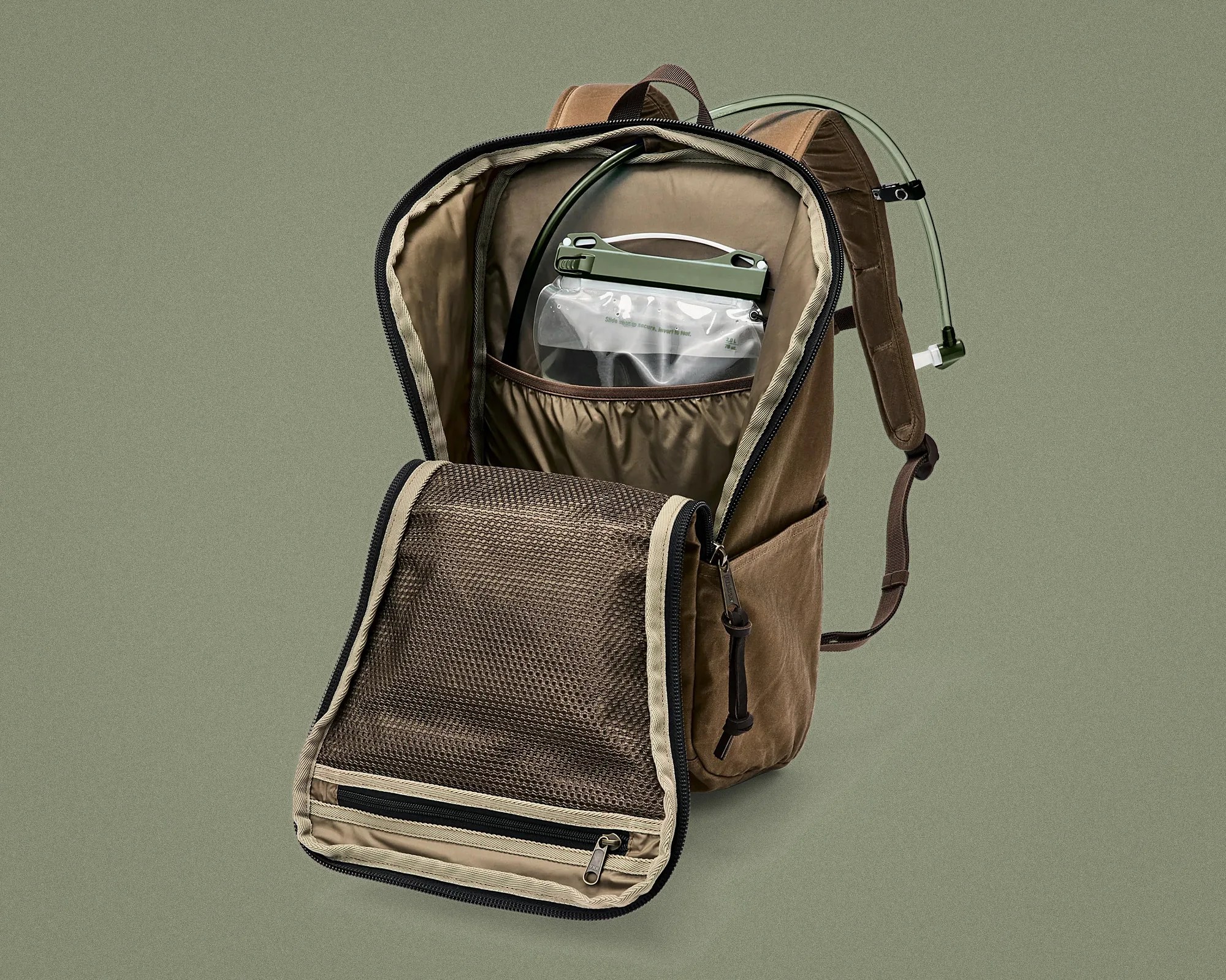 Brown backpack with hydration bladder and mesh zippered pocket inside, shown open against a green background.
