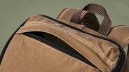 Close-up of a brown canvas backpack with a partially open black zipper pocket and a brown woven handle.