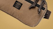 Brown canvas Filson pouch with black zipper and leather pull tabs on a tan background.