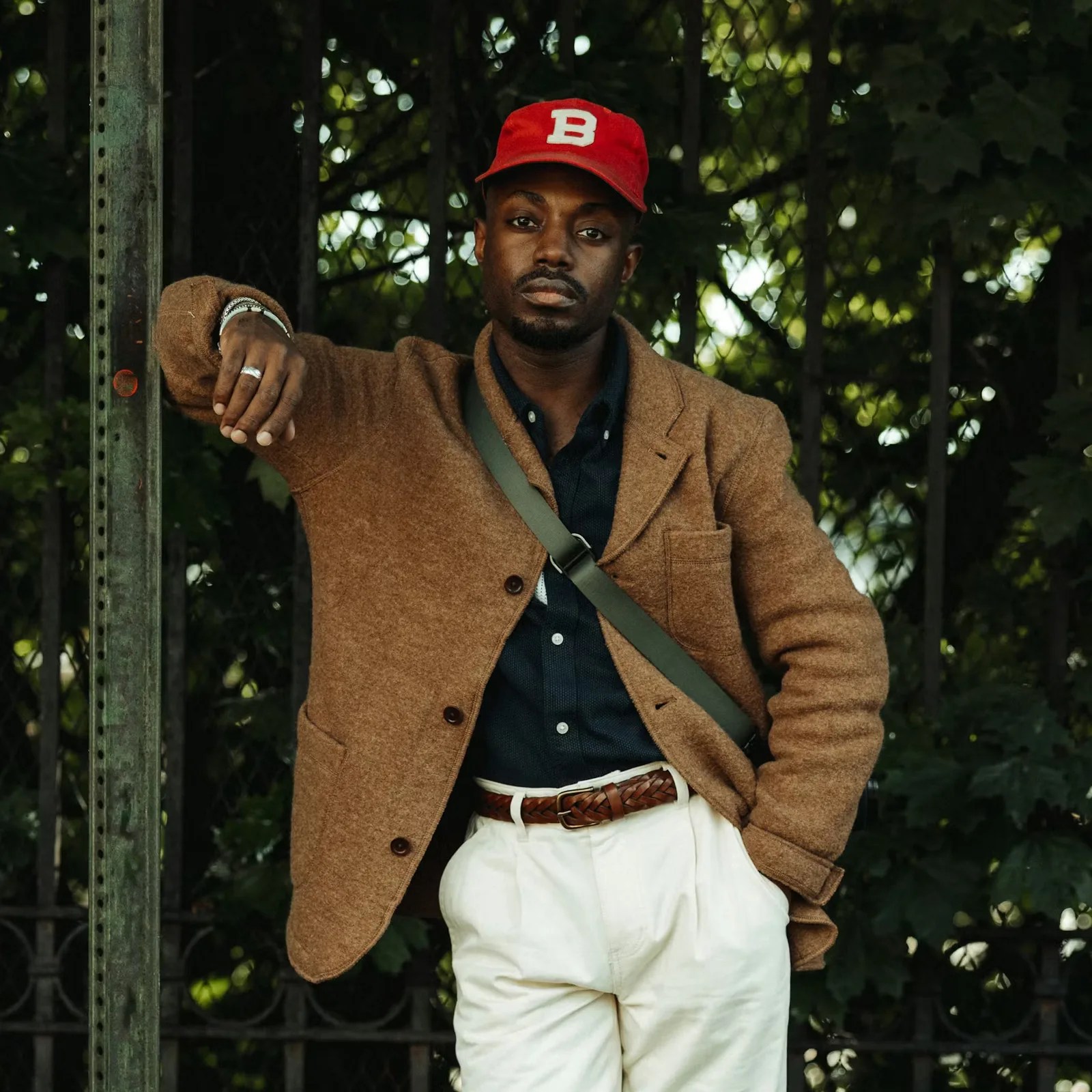 Man wearing a brown wool blazer, navy shirt, white pants with a braided brown belt, red cap with a white "B," and green crossbody bag.