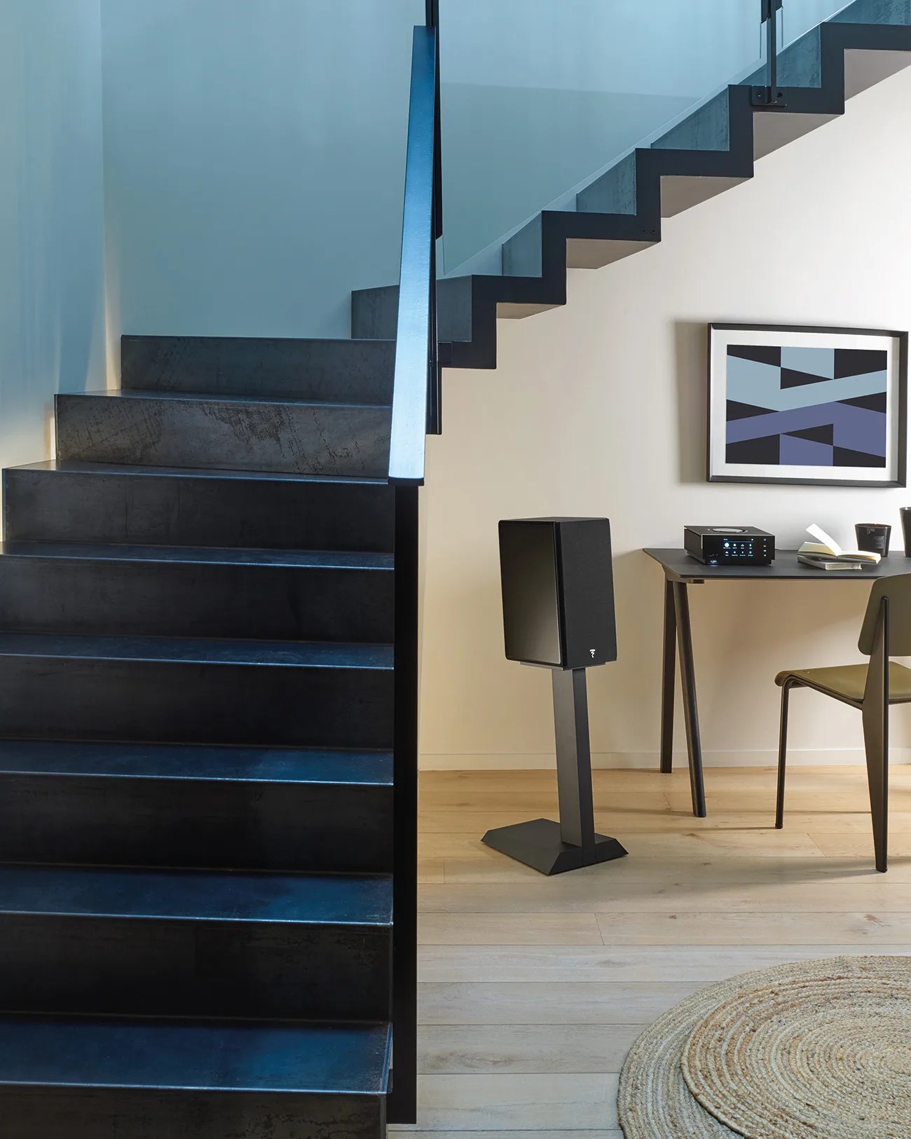 Black metal staircase next to a modern workspace with a black speaker on a stand, a desk, chair, and abstract wall art.