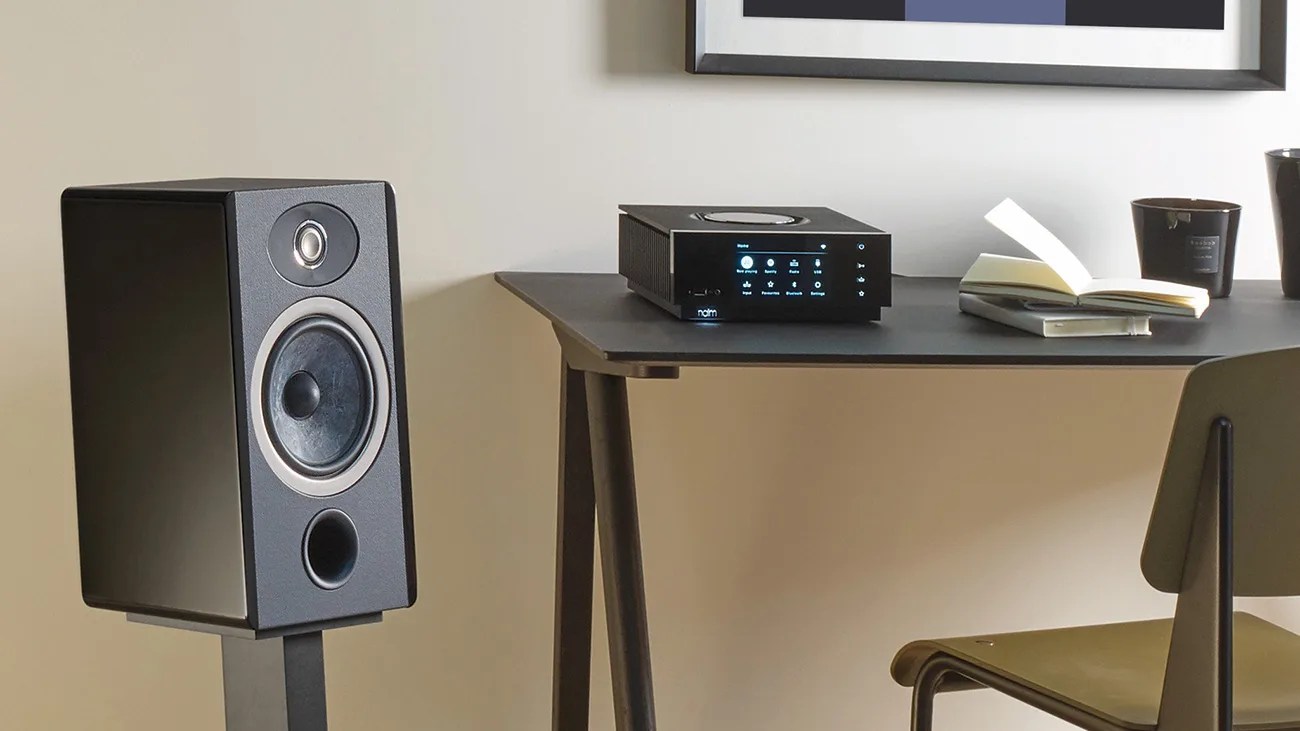 Black bookshelf speaker on a stand next to a black audio player with a digital display on a desk.
