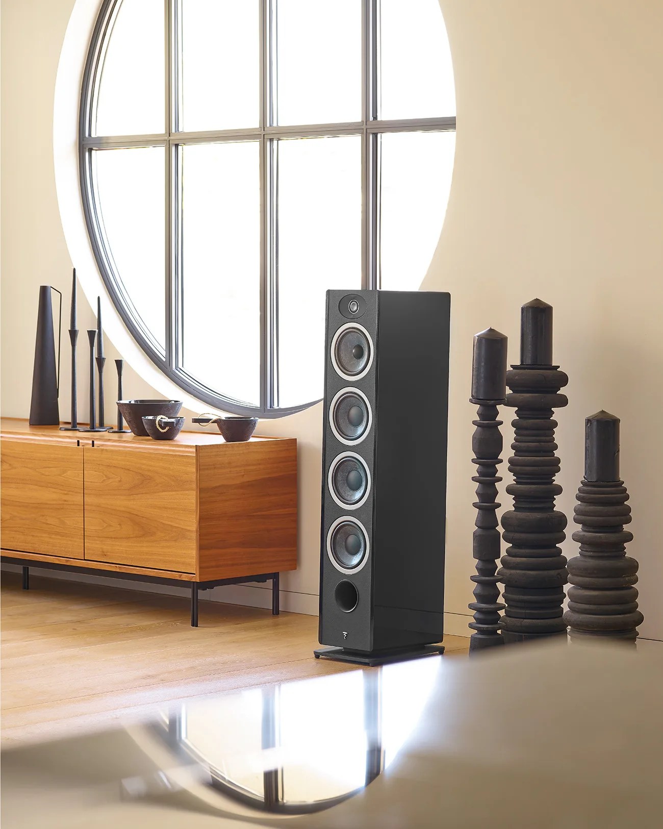 Tall black floor-standing speaker with four drivers next to a wooden sideboard and sculptural black candles.