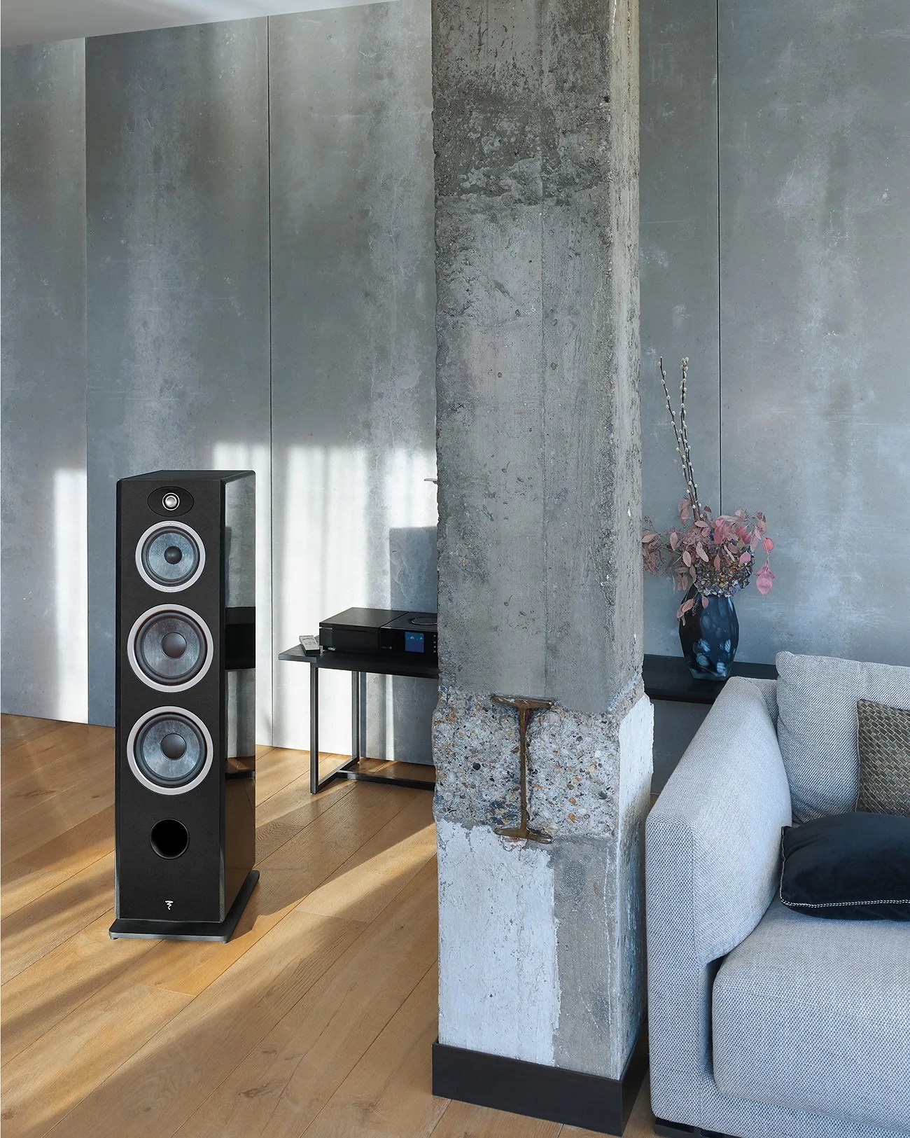 Black floor-standing speaker with three silver-ringed drivers on a wooden floor next to a concrete pillar and gray sofa.