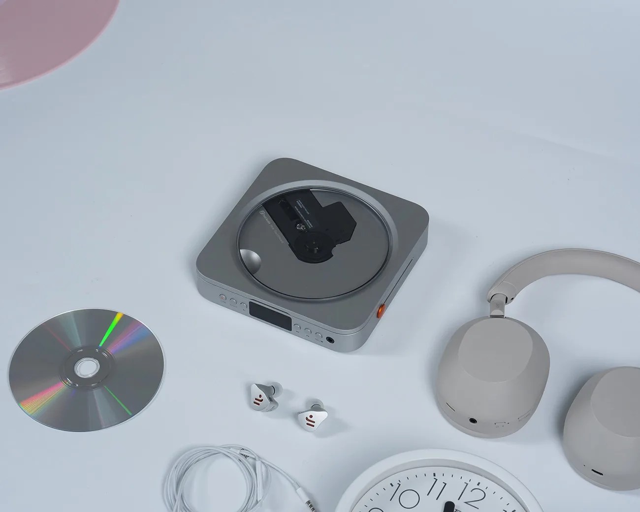 Silver portable CD player with a CD inside, silver CD disc, white wired earphones, beige over-ear headphones, and a white clock on a light surface.