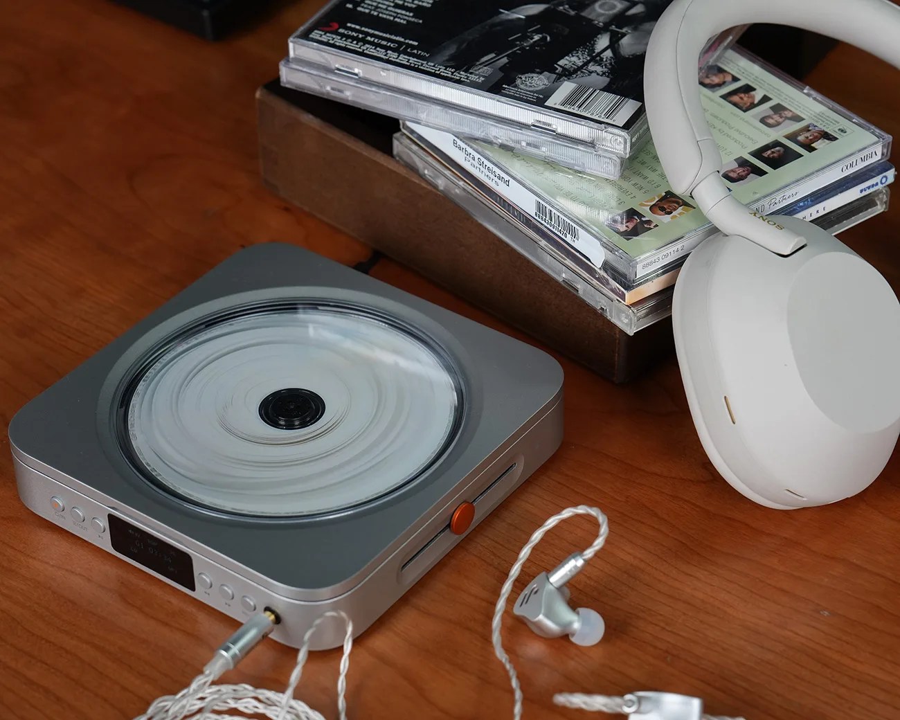 Silver portable CD player with a clear lid, silver wired earphones, white over-ear headphones, and a wooden box with stacked CD cases on a wooden surface.