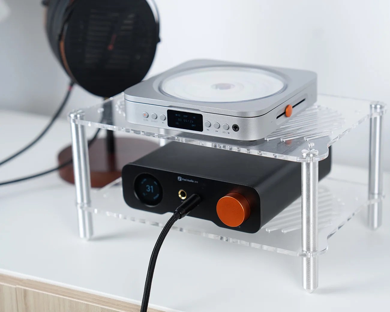 Clear acrylic two-tier audio equipment stand holding a silver CD player and a black audio amplifier with orange knobs.