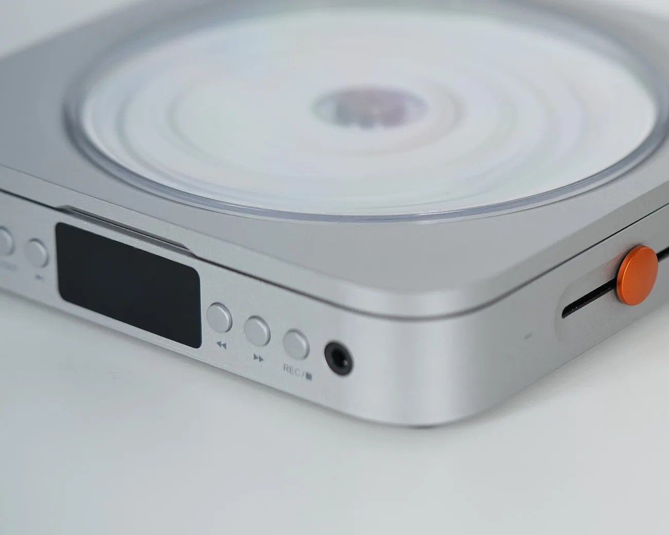 Silver portable CD player with black display screen and orange volume knob on a white surface.