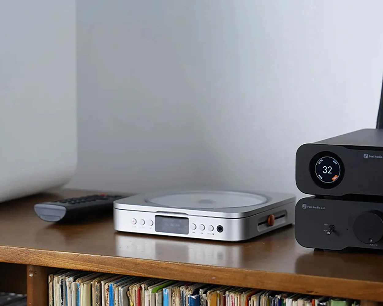 Silver compact turntable with buttons and display on a wooden shelf next to black audio equipment and a remote control.
