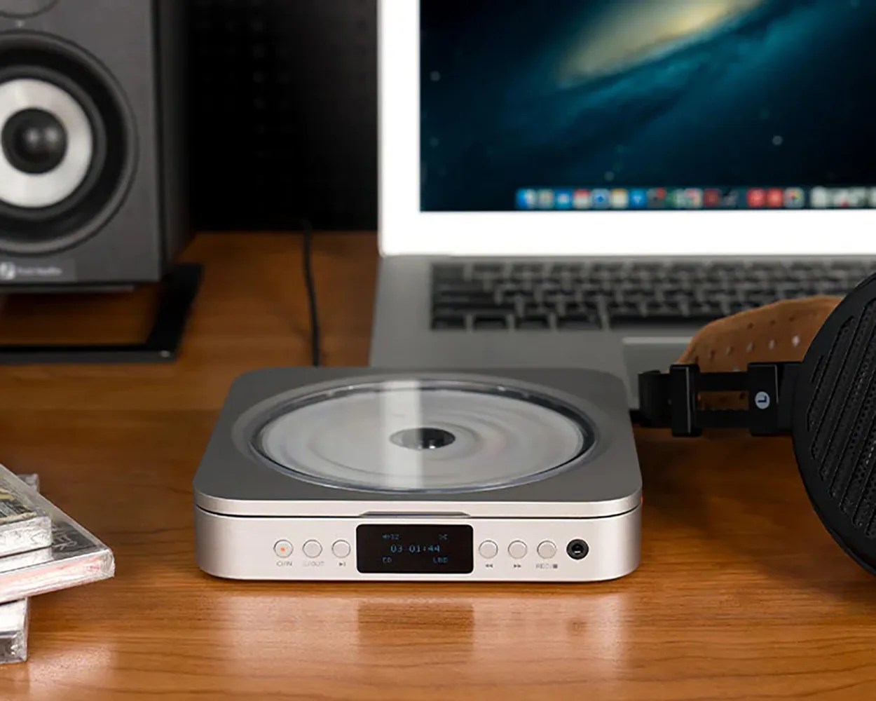 Silver compact disc player with digital display on a wooden desk next to a laptop and black headphones.