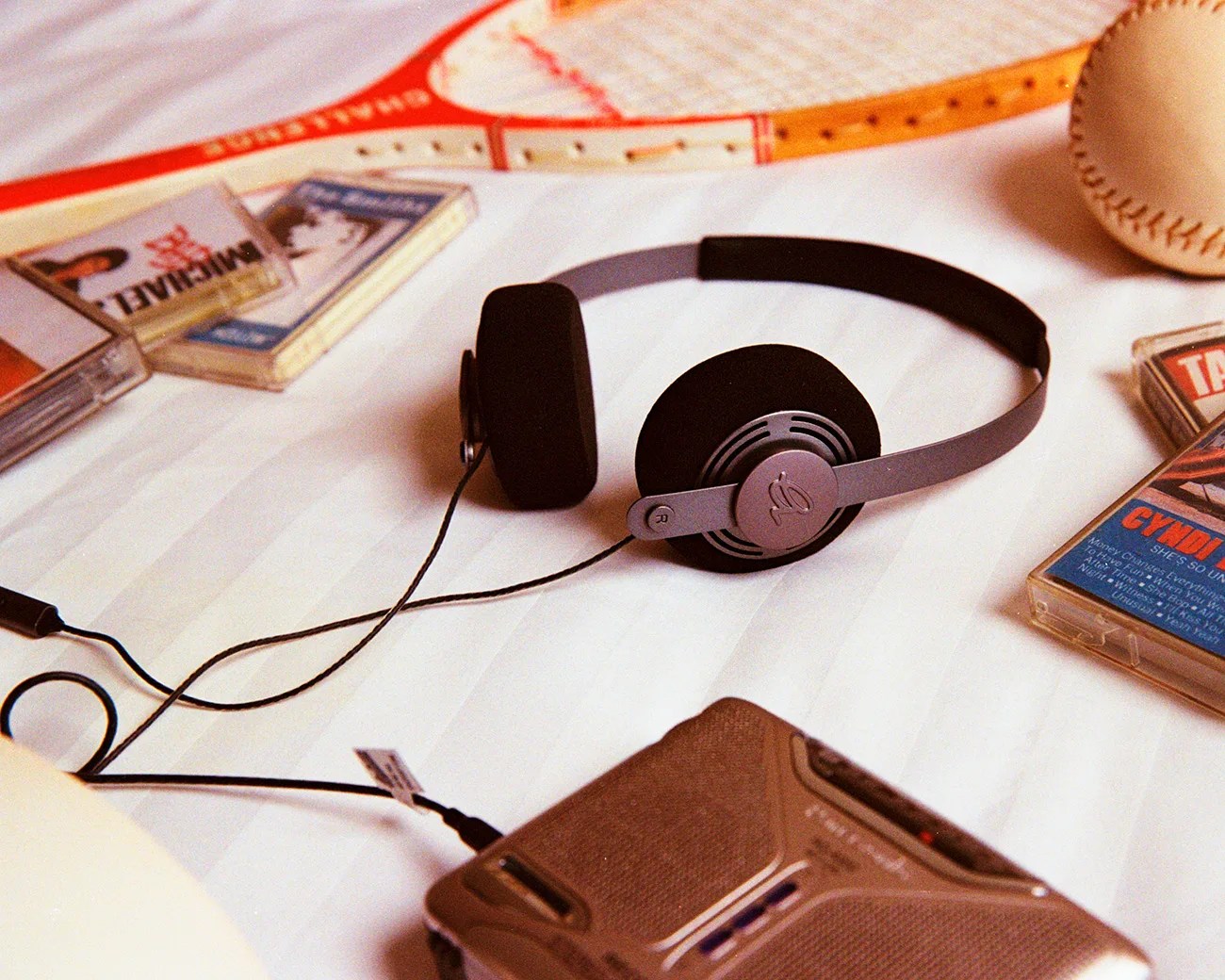 Black and gray wired headphones connected to a silver cassette player on a white surface with cassette tapes and a baseball.