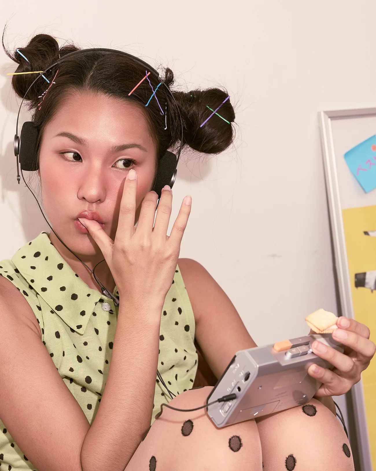Young person with colorful hairpins in double buns wearing headphones and a green polka-dot sleeveless top, holding a portable cassette player and a cracker.