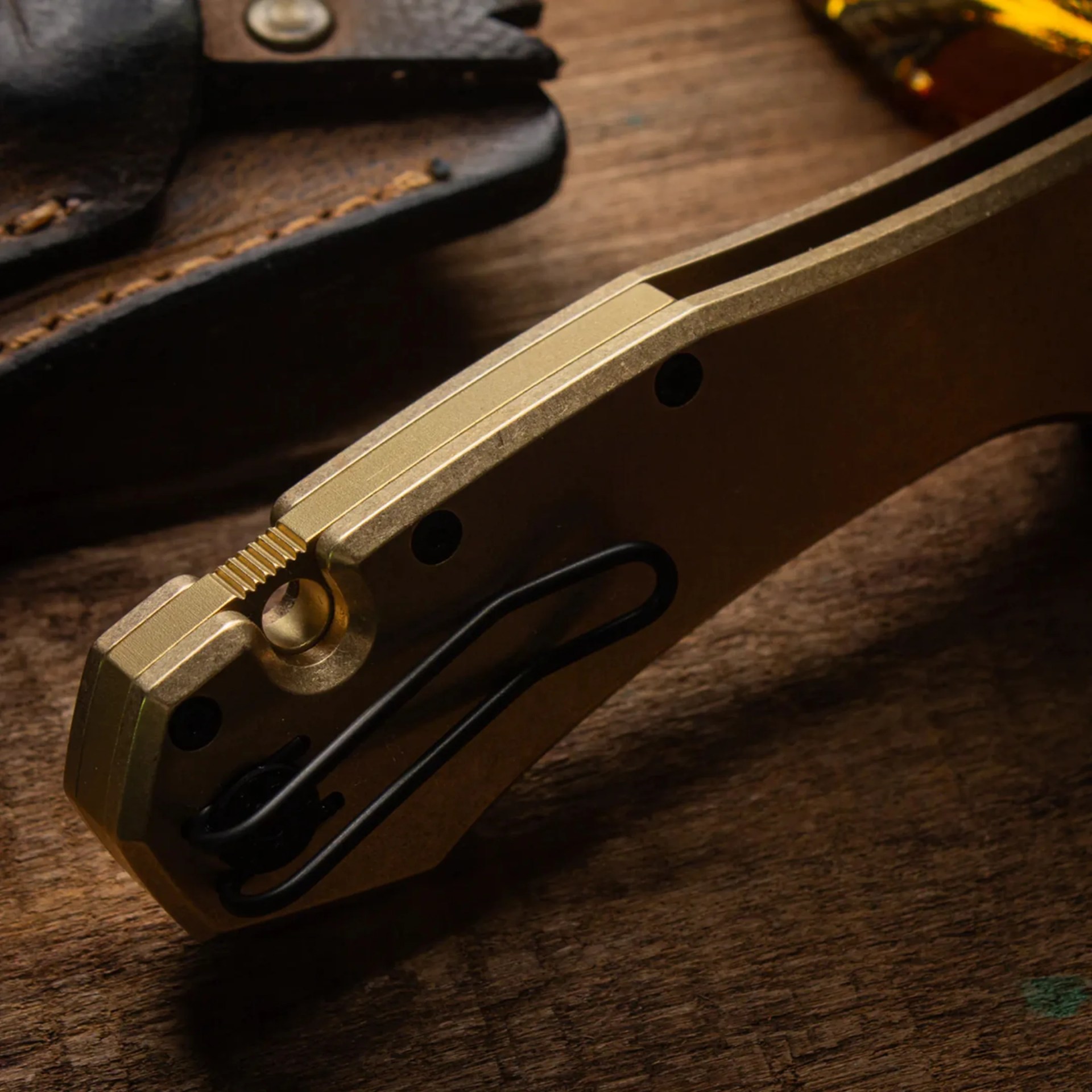Close-up of a bronze-colored folding knife handle with a black metal clip on a wooden surface.