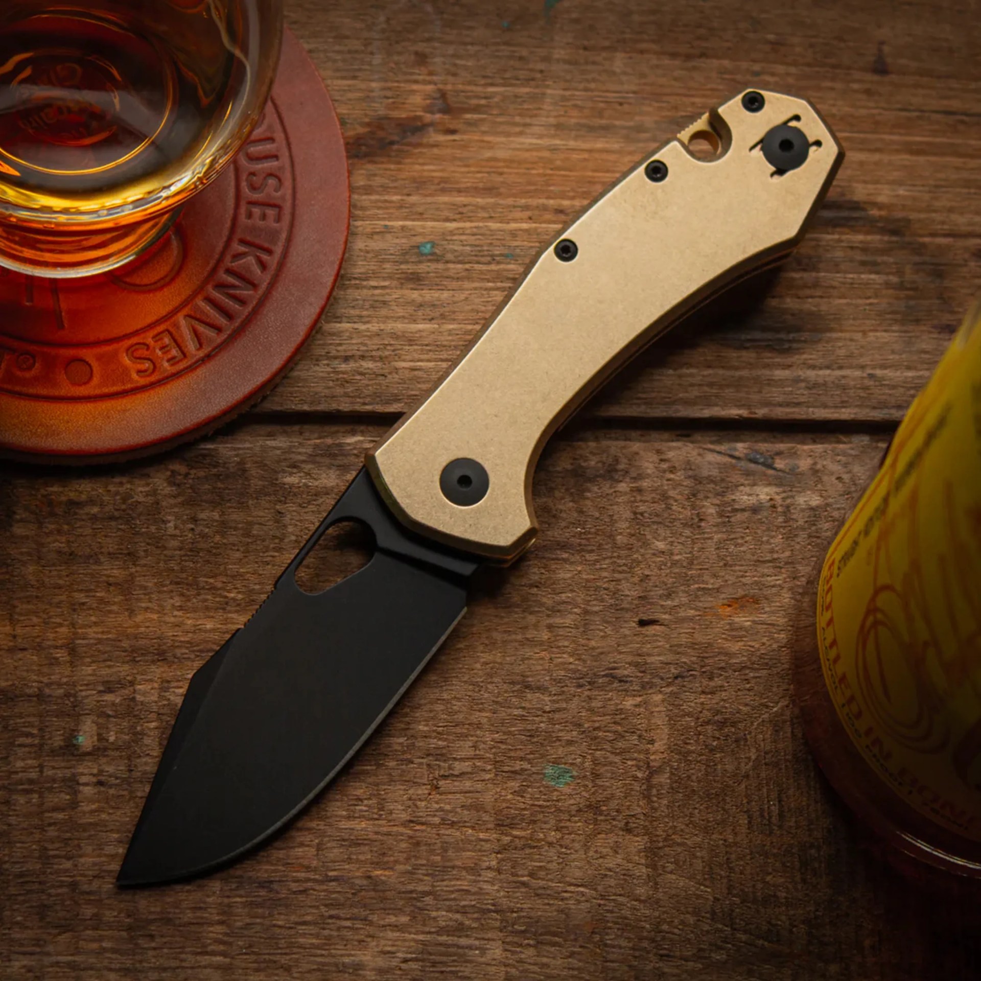 Folding knife with a black blade and gold handle on a wooden surface next to a glass and a bottle.