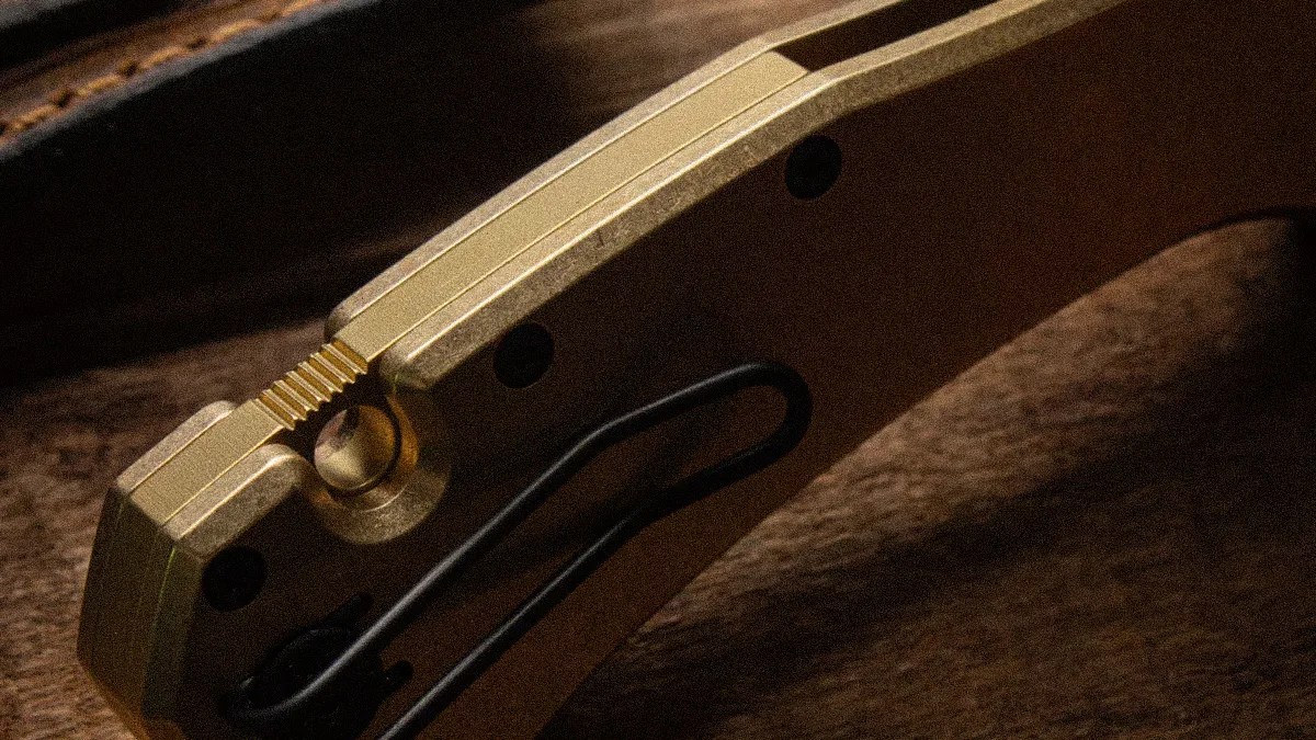 Close-up of a gold-colored metal folding knife handle with a black clip on a brown textured surface.