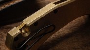 Close-up of a gold-colored metal folding knife handle with a black clip on a brown textured surface.