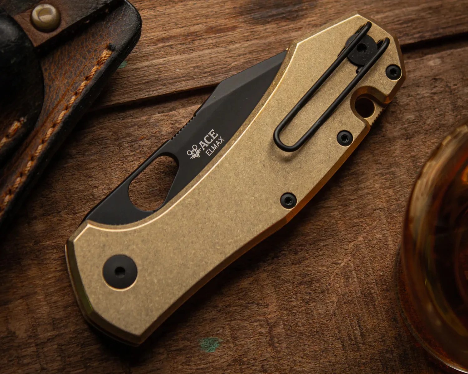 Folded pocket knife with a gold-toned handle and black blade marked "ACE ELMAX" on a wooden surface.