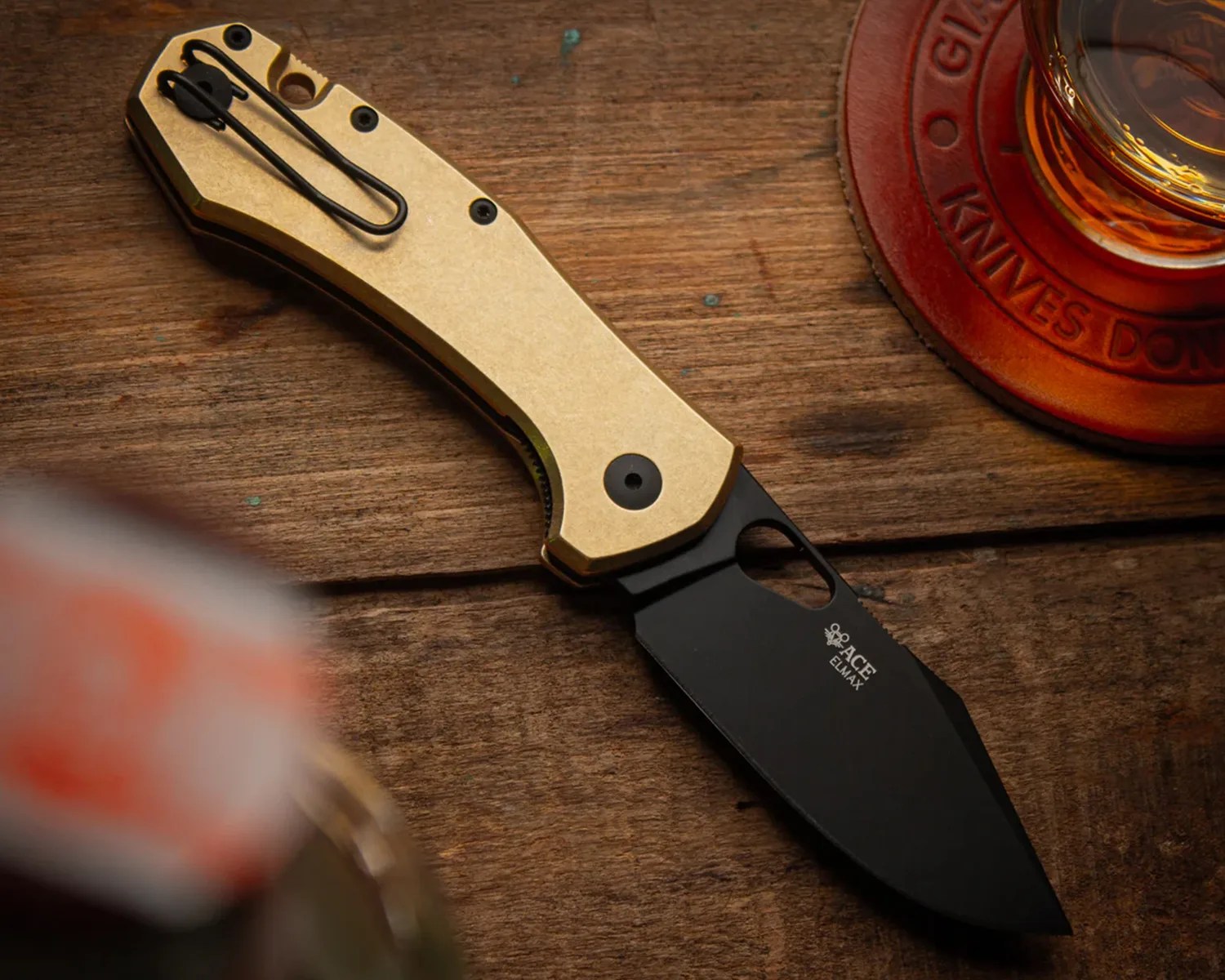 Folding knife with a gold handle and black blade labeled "ACE ELMAX" on a wooden surface.