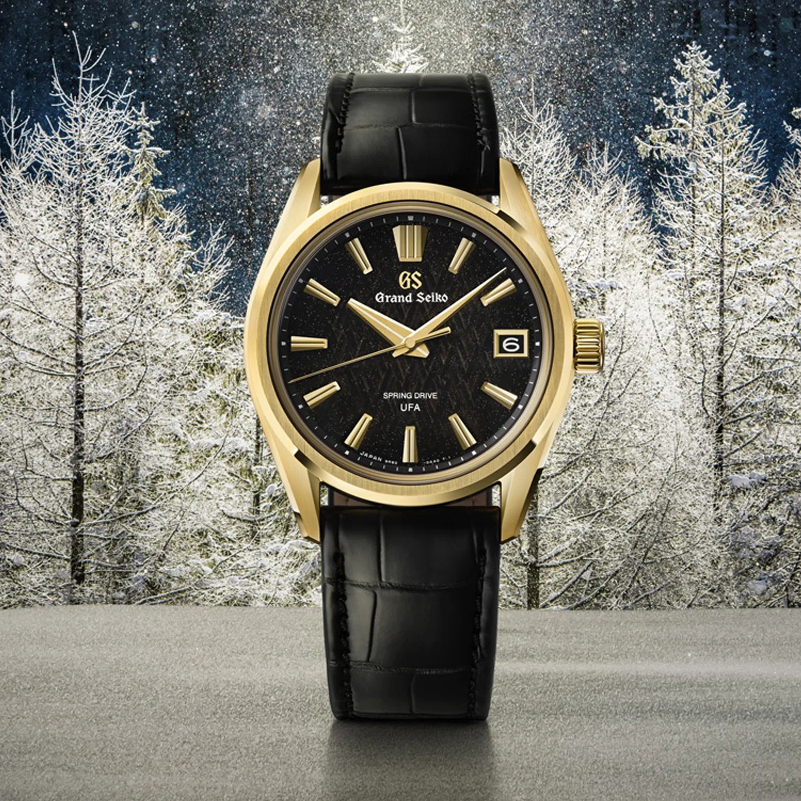 Gold Grand Seiko wristwatch with black textured dial and black leather strap against snowy forest background.