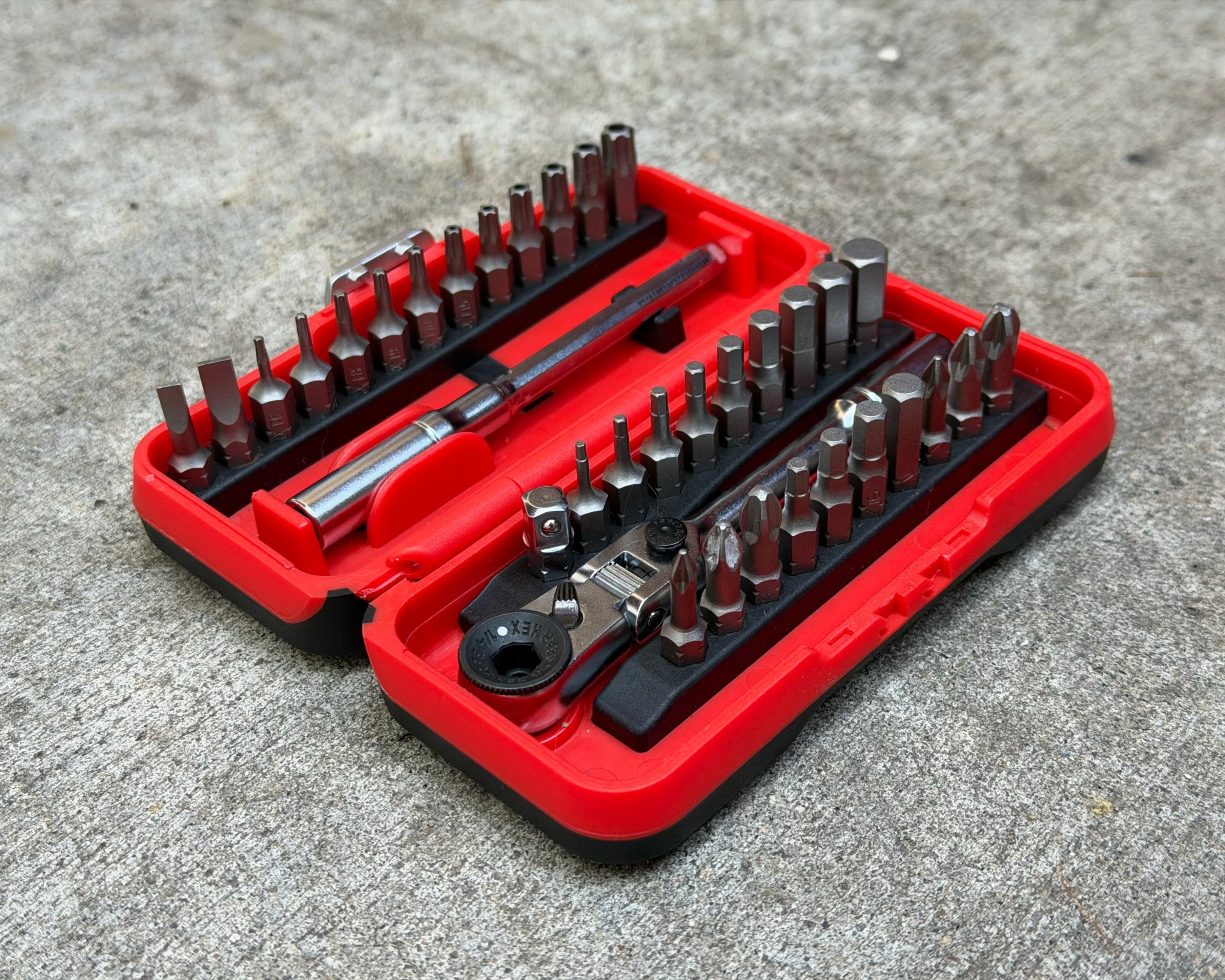 Red and black plastic case with a set of various metal screwdriver bits and a ratchet wrench on a concrete surface.