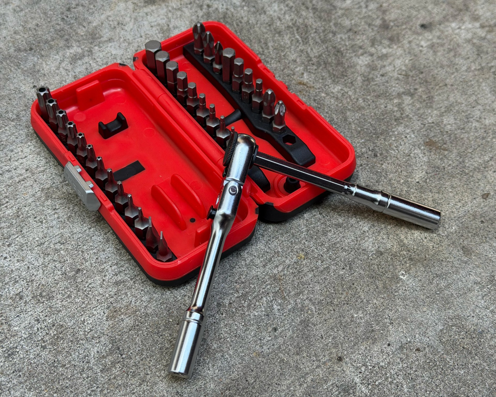 Red plastic case with various metal screwdriver bits and a ratchet wrench on a concrete surface.