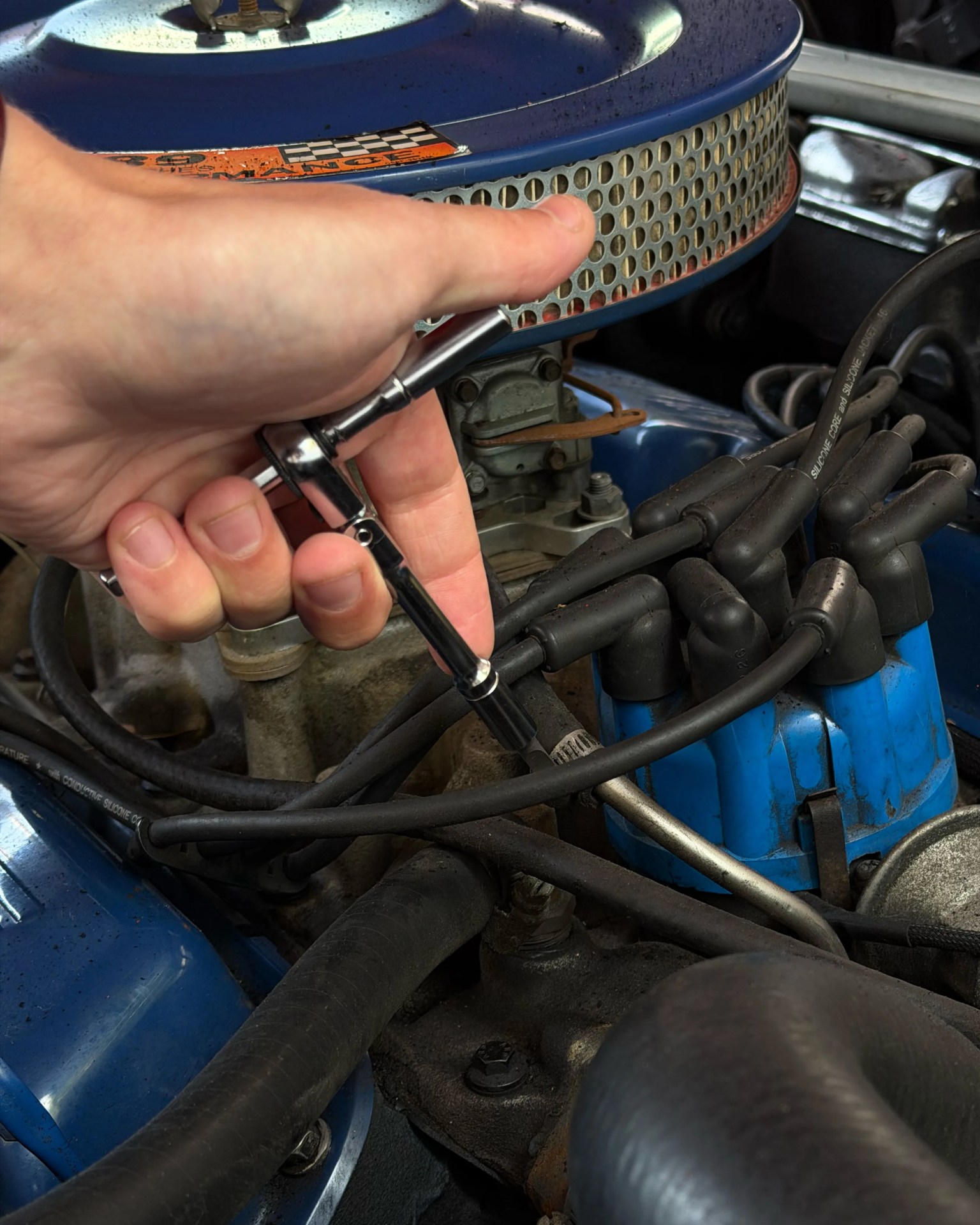 Hand holding a ratchet wrench working on a blue engine with black ignition wires and a round air filter.