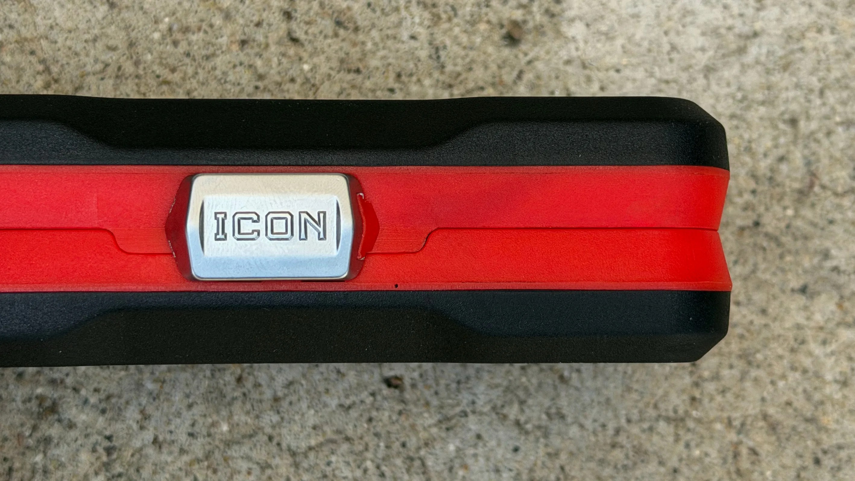 Close-up of a black and red tool handle with a silver button labeled "ICON" on a concrete surface.