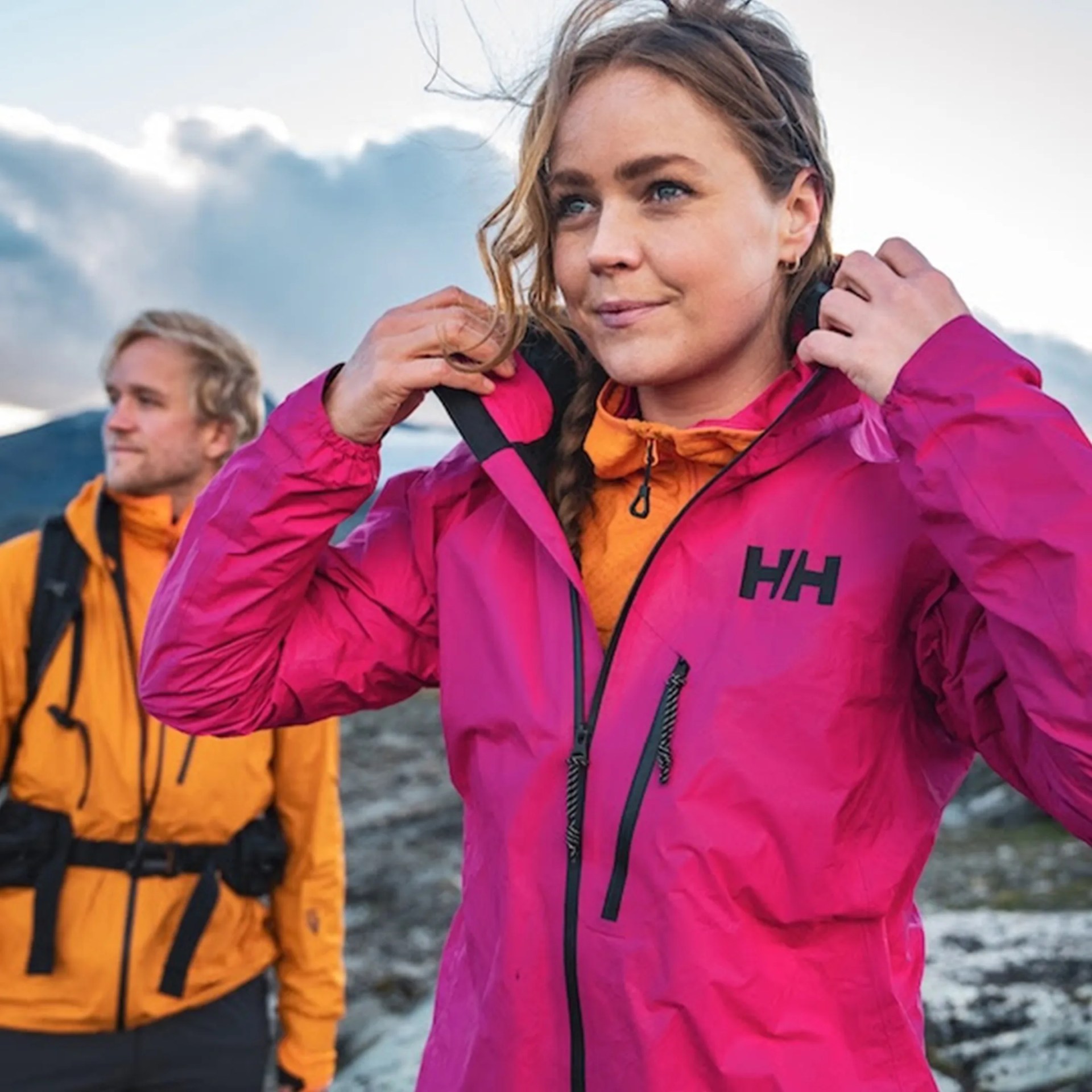Woman adjusting the hood of a bright pink Helly Hansen jacket with a man in an orange jacket in the background outdoors.