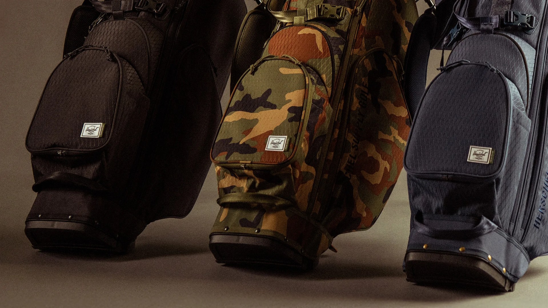 Three Herschel golf bags in black, camouflage, and navy colors standing on a brown surface.