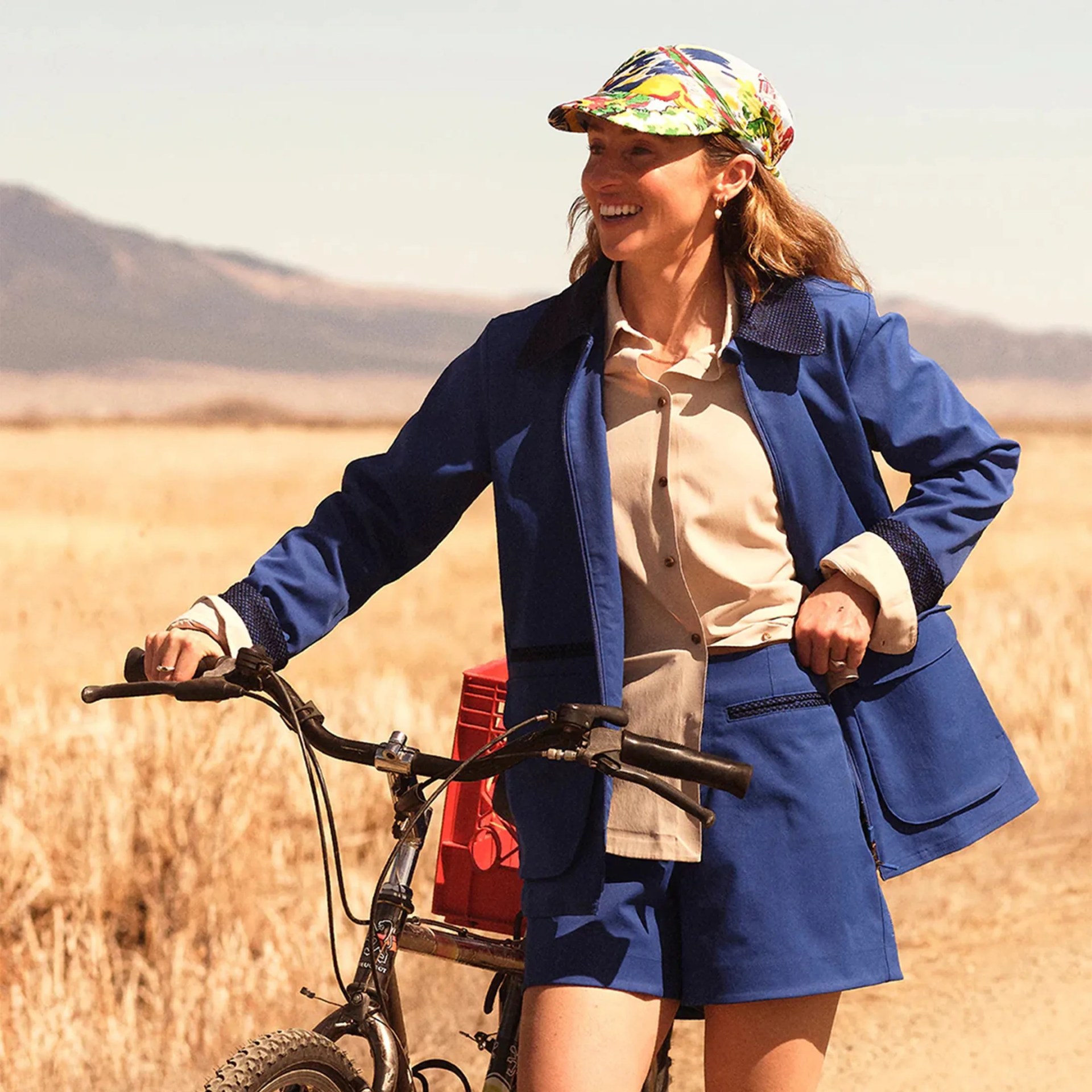 Person wearing a blue jacket and shorts with a beige shirt and colorful cap, holding a bicycle in a dry field.